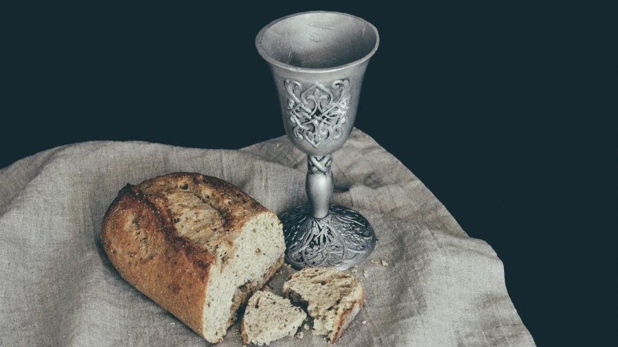 A silver chalice and a broken loaf of bread on a beige cloth.